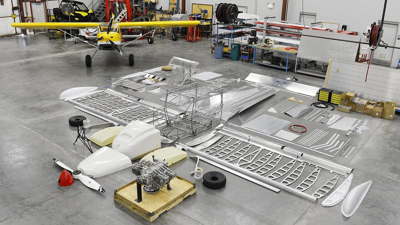 An aircraft kit laid out on a hangar floor, with a finished aircraft of the same kind behind it